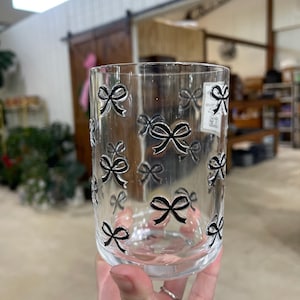 May include: A clear glass tumbler adorned with black bow designs. The bows are evenly spaced around the glass, creating a decorative pattern. The glass is held in a person's hand, with the background slightly blurred.