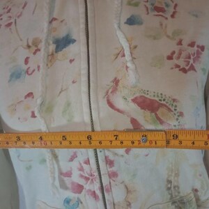 May include: Cream-colored zip-up hoodie with a floral pattern in pink, red, blue, and green. The hoodie has a drawstring hood and a front zipper. The measurement across the chest is approximately 10 inches.