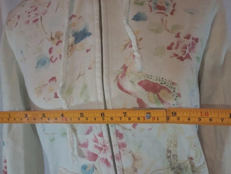 May include: Cream-colored zip-up hoodie with a floral pattern in pink, red, blue, and green. The hoodie has a drawstring hood and a front zipper. The measurement across the chest is approximately 10 inches.