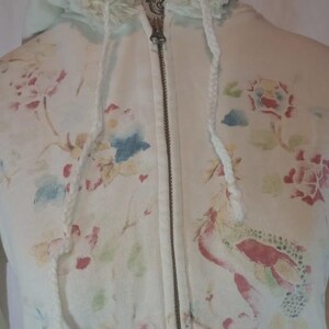 May include: Cream-colored zip-up hoodie with a floral pattern in shades of pink, red, blue, and green. The hoodie features a drawstring hood with fringe and a silver zipper. The overall design has a vintage, watercolor aesthetic.