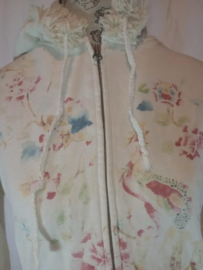 May include: Cream-colored zip-up hoodie with a floral pattern in shades of pink, red, blue, and green. The hoodie features a drawstring hood with fringe and a silver zipper. The overall design has a vintage, watercolor aesthetic.