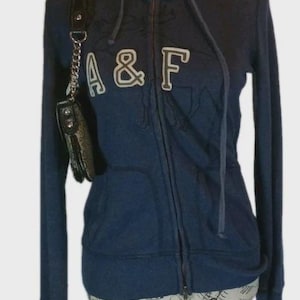 May include: Navy blue zip-up hoodie with the letters "A & F" in white. The hoodie has a drawstring hood and front pockets. A small black purse with a silver chain hangs on the left side. A silver necklace is around the neck.