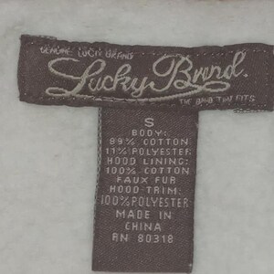 May include: Close-up of a brown Lucky Brand tag with white text on a white and pink background. The tag reads "GENUINE LUCKY BRAND" and "THE BAND THAT FITS". The tag also lists the fabric content and "MADE IN CHINA".