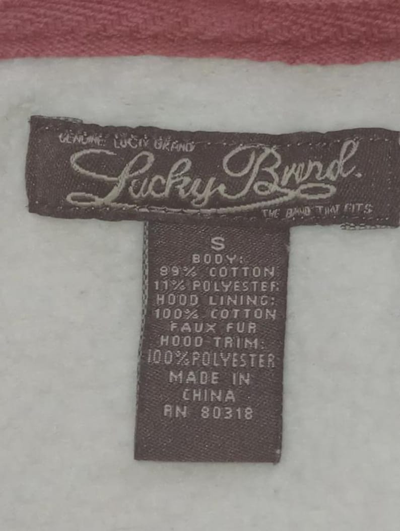 May include: Close-up of a brown Lucky Brand tag with white text on a white and pink background. The tag reads "GENUINE LUCKY BRAND" and "THE BAND THAT FITS". The tag also lists the fabric content and "MADE IN CHINA".