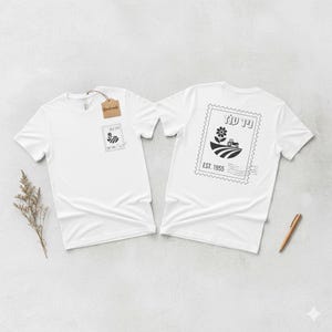 May include: Two white t-shirts with a black and white graphic design. The front of the shirt has a small stamp design, and the back has a larger stamp design with a flower, tractor, and the text "EST. 1955". A small brown tag hangs from one shirt.