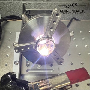 May include: A close-up of a metal rotary table with a bright spark of light in the center. The table is mounted on a base with a clamp attached to the side. The text "ADIRONDACK INLAY STUDIO" is visible in the upper right corner of the image.