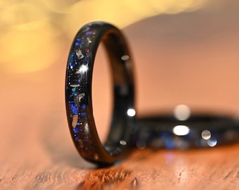 May include: Two black wedding bands with blue and silver inlay. The bands are on a wooden surface.