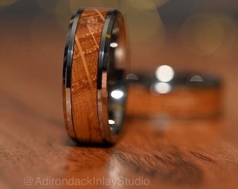 May include: A close-up of a wooden and stainless steel wedding band. The band has a brown wood inlay with a silver metal border. The band is lying on a wooden surface.
