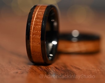 May include: A black tungsten carbide ring with a brown wood inlay and a thin gold line. The ring is on a wooden surface.