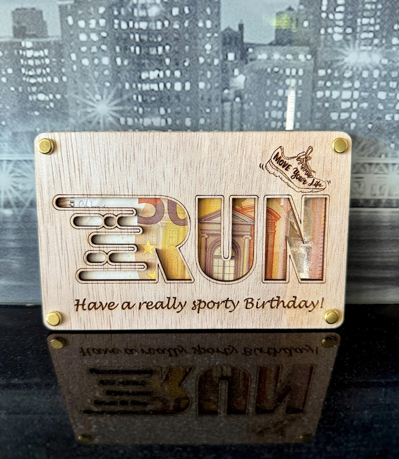 May include: Wooden money box with the word "RUN" cut out, revealing Euro banknotes. The box has the phrase "Have a really sporty Birthday!" and a running shoe graphic with the text "Move Your Life". Gold-colored screws secure the box.