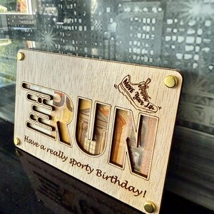 May include: A wooden gift card holder with the word "RUN" cut out, revealing currency inside. The design includes a running shoe graphic and the phrase "Move Your Life." The bottom text reads "Have a really sporty Birthday!"