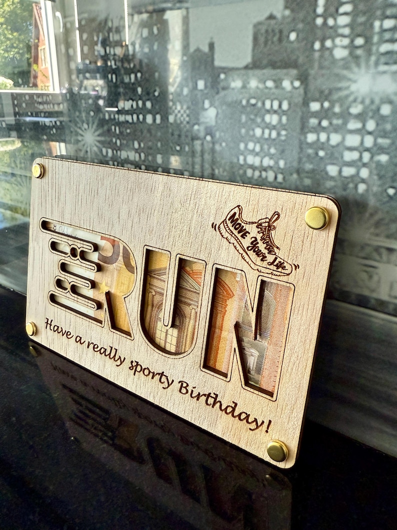 May include: A wooden gift card holder with the word "RUN" cut out, revealing currency inside. The design includes a running shoe graphic and the phrase "Move Your Life." The bottom text reads "Have a really sporty Birthday!"