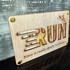 May include: Wooden plaque with the word "RUN" cut out, revealing currency inside. The plaque has the text "Have a really sporty Birthday!" and a small shoe graphic. Gold-colored rivets secure the corners.