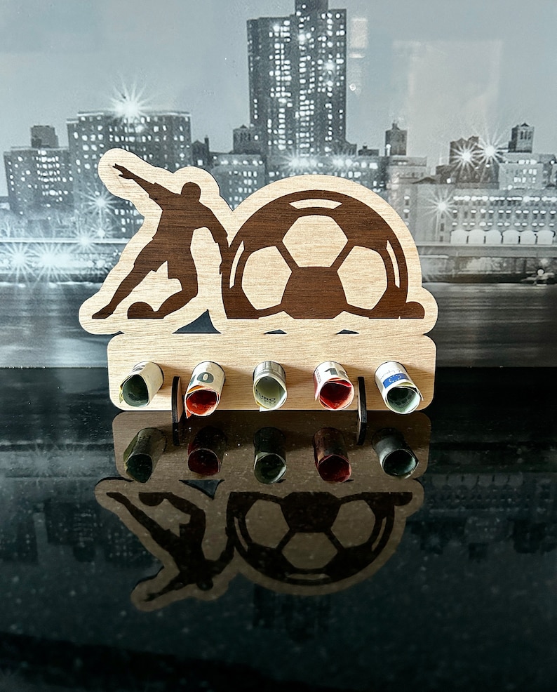 Pode incluir: Porta-dinheiro de madeira em forma de jogador de futebol chutando uma bola. O porta-dinheiro tem seis ranhuras para notas enroladas.