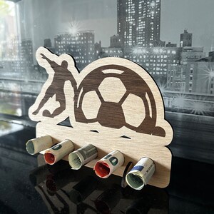 Pode incluir: Porta-dinheiro de madeira em forma de jogador de futebol chutando uma bola. O porta-dinheiro tem cinco ranhuras para notas enroladas.