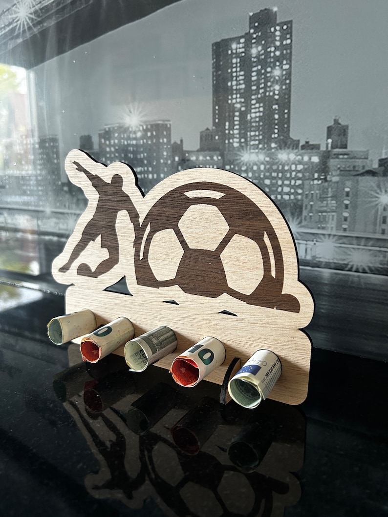 Pode incluir: Porta-dinheiro de madeira em forma de jogador de futebol chutando uma bola. O porta-dinheiro tem cinco ranhuras para notas enroladas.
