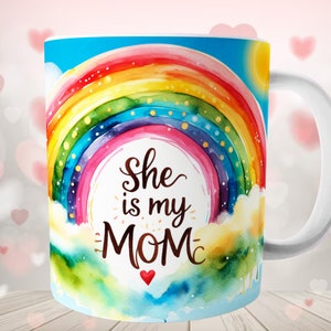 Pode incluir: Caneca de cerâmica branca com um design de arco-íris colorido e o texto "She is my MOM" em letras marrons. O arco-íris é pintado em estilo aquarela sobre um fundo de céu azul.
