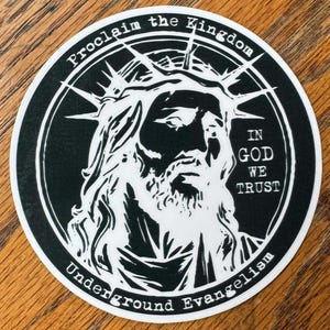 May include: Black and white sticker with a stylized image of Jesus wearing a crown of thorns. The text "Proclaim the Kingdom" is at the top of the sticker, and "In God We Trust" is on the right side. The text "Underground Evangelism" is at the bottom of the sticker.