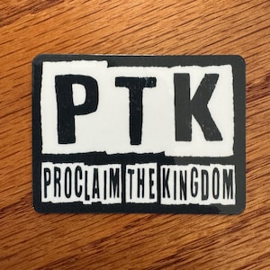 May include: Rectangular sticker with the letters "PTK" in bold, black font on a white background. Below, the words "PROCLAIM THE KINGDOM" are printed in a similar style. The sticker has a black border and is set against a wooden surface.