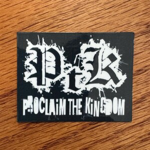 May include: A rectangular black sticker with white text and a distressed design. The sticker features the letters "DTK" in a gothic font, with the words "PROCLAIM THE KINGDOM" below.