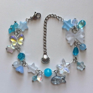May include: A silver-toned metal charm bracelet featuring an assortment of blue, white, and iridescent beads. The charms include stars, a butterfly, a bow, a fish, and a seashell pendant attached to a central chain link with a lobster clasp.