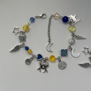 May include: A silver charm bracelet with a variety of charms, including stars, moons, hearts, wings, a spider, a skull, and a spiral. The bracelet also features yellow and blue beads.