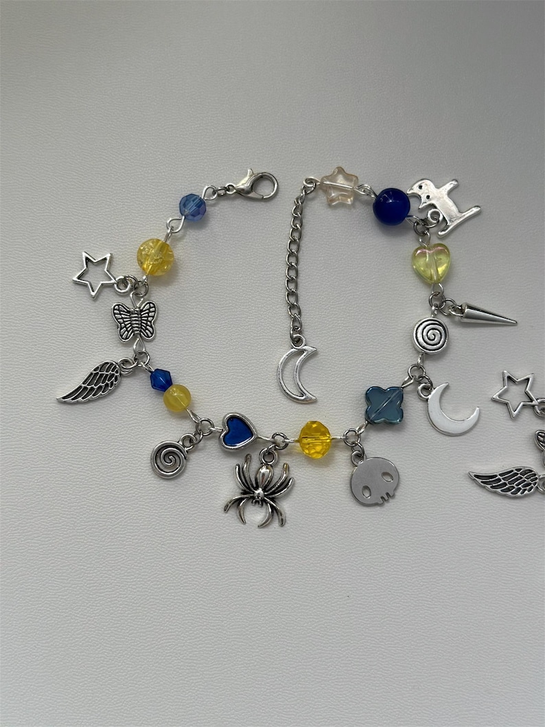 May include: A silver charm bracelet with a variety of charms, including stars, moons, hearts, wings, a spider, a skull, and a spiral. The bracelet also features yellow and blue beads.