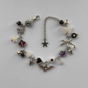 May include: A silver-toned charm bracelet featuring various charms, including stars, butterflies, hearts, and a bat. The bracelet has a lobster clasp closure and a chain extender. The charms are in black, white, silver, and purple.