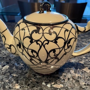 May include: Cream-colored ceramic teapot with a black, ornate, swirling pattern. The teapot has a curved spout, a rounded handle, and a lid with a small, dark-colored knob. The rim and spout are trimmed with a dark brown color.