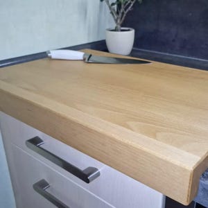 Könnte beinhalten: Ein helles Holzschneidebrett mit einem Edelstahlmesser und weißem Griff. Das Brett befindet sich auf einem weißen Schrank mit silbernen Griffen. Im Hintergrund befindet sich eine kleine Topfpflanze.