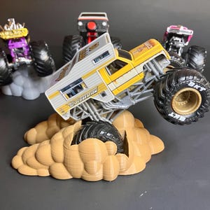 May include: A yellow and white monster truck with the words "Wreckreation" on the side, driving over a brown cloud-shaped base. The truck has large black tires with gold rims. There are other monster trucks in the background.