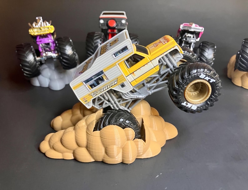 May include: A yellow and white monster truck with the words "Wreckreation" on the side, driving over a brown cloud-shaped base. The truck has large black tires with gold rims. There are other monster trucks in the background.