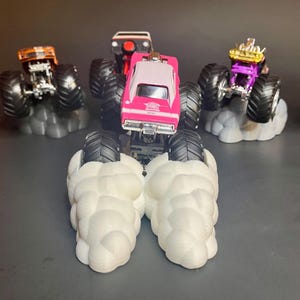 May include: A pink monster truck with black wheels and a white roof, with smoke coming out from under the wheels. The truck is on a white cloud-shaped base. There are other monster trucks in the background.