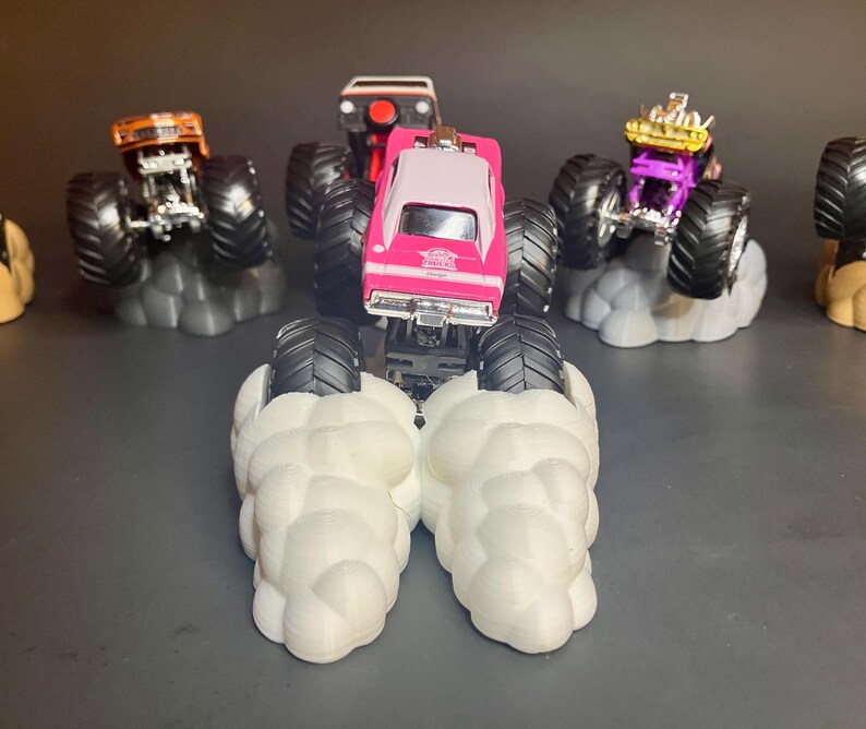 May include: A pink monster truck with black wheels and a white roof, with smoke coming out from under the wheels. The truck is on a white cloud-shaped base. There are other monster trucks in the background.