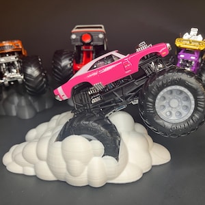 May include: A pink monster truck with a white stripe, large black wheels, and a black chassis, is driving over a white cloud-shaped base. Three other monster trucks are in the background, one orange, one purple, and one white.