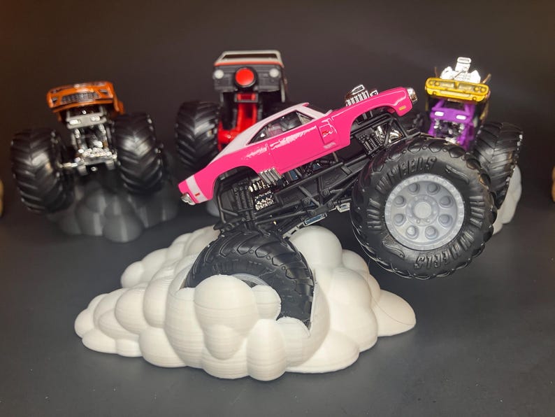 May include: A pink monster truck with a white stripe, large black wheels, and a black chassis, is driving over a white cloud-shaped base. Three other monster trucks are in the background, one orange, one purple, and one white.