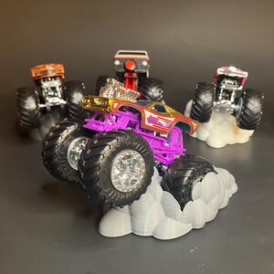 May include: A purple monster truck with gold accents and a red and white logo on the side. The truck is on a gray cloud-shaped base. There are other monster trucks in the background.