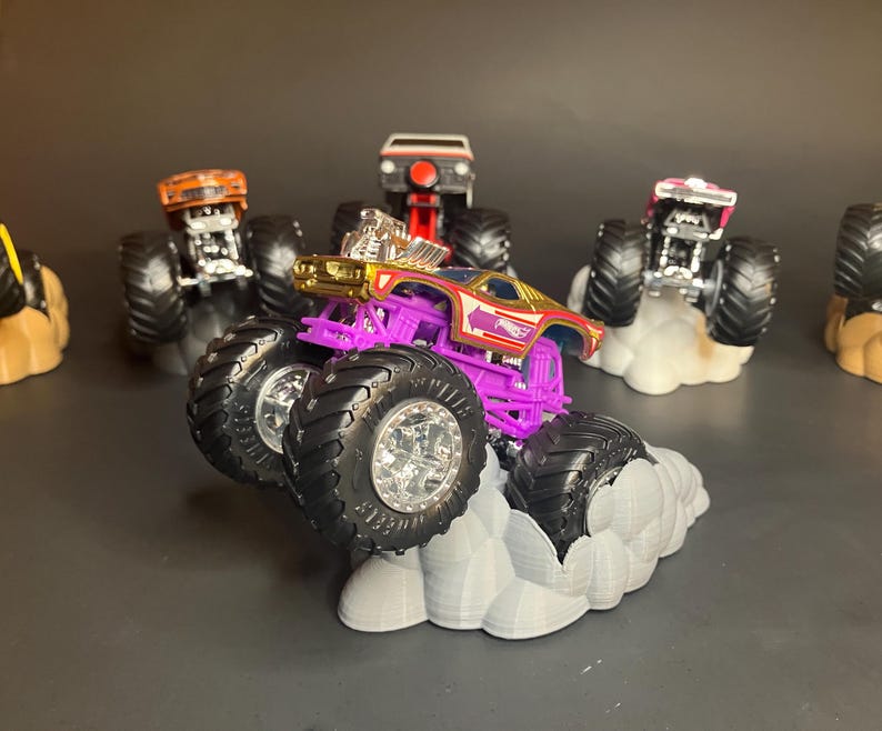 May include: A purple monster truck with gold accents and a red and white logo on the side. The truck is on a gray cloud-shaped base. There are other monster trucks in the background.
