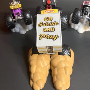 May include: A yellow monster truck with a white sign that says "GO Outside AND Play". The truck is on a brown, textured base. There are two other monster trucks in the background.