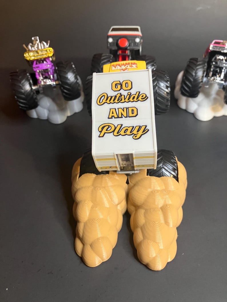 May include: A yellow monster truck with a white sign that says "GO Outside AND Play". The truck is on a brown, textured base. There are two other monster trucks in the background.