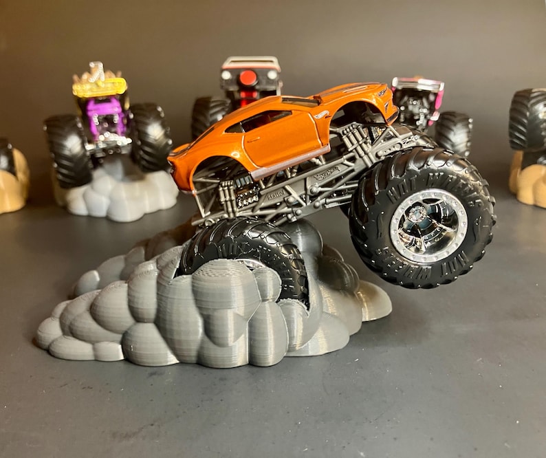 May include: Orange monster truck with large black wheels and a gray cloud base. The truck is a modified version of a Chevrolet Camaro.