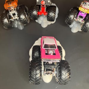 May include: Four monster trucks with large tires and different paint schemes. The trucks are on a white cloud-shaped base. The trucks are a pink Dodge Charger, a white van, a gold and black car, and an orange Camaro.