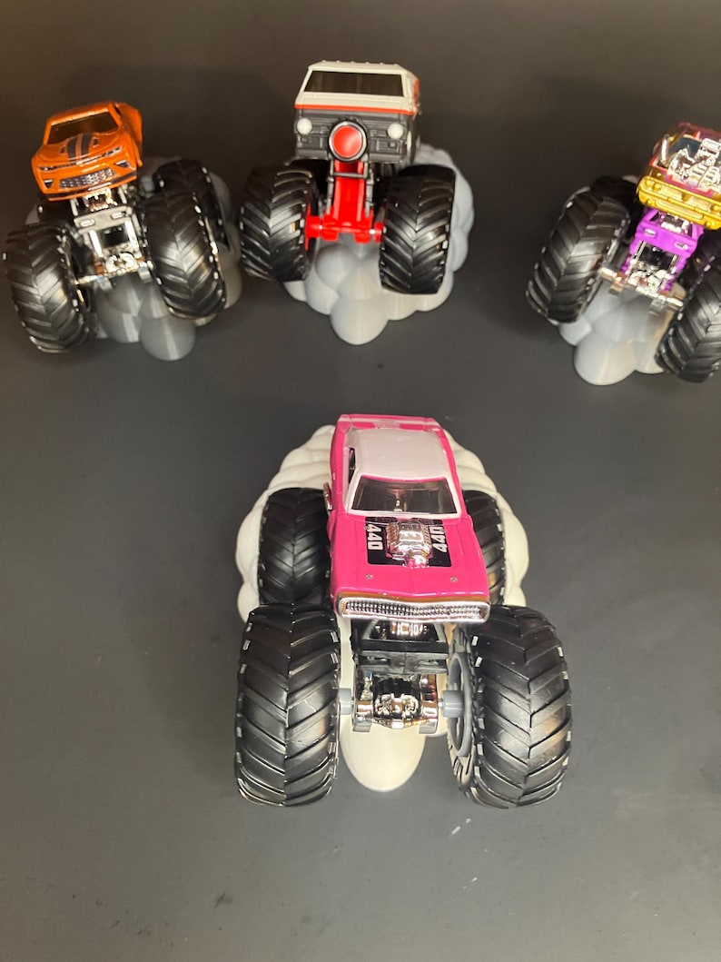May include: Four monster trucks with large tires and different paint schemes. The trucks are on a white cloud-shaped base. The trucks are a pink Dodge Charger, a white van, a gold and black car, and an orange Camaro.