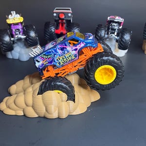 May include: A blue and orange monster truck toy with the words "Grave Digger" on the side, driving up a brown cloud-shaped ramp. The truck has yellow wheels and is surrounded by other monster trucks.