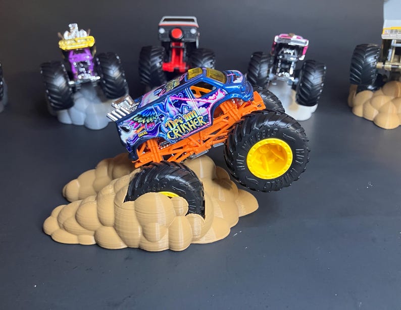 May include: A blue and orange monster truck toy with the words "Grave Digger" on the side, driving up a brown cloud-shaped ramp. The truck has yellow wheels and is surrounded by other monster trucks.