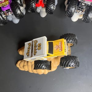 May include: A yellow monster truck toy with a white box on top that says "GO OUTSIDE AND PLAY". The truck has black wheels and a black star on the side. The truck is sitting on a brown surface.