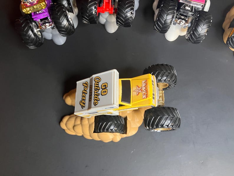 May include: A yellow monster truck toy with a white box on top that says "GO OUTSIDE AND PLAY". The truck has black wheels and a black star on the side. The truck is sitting on a brown surface.