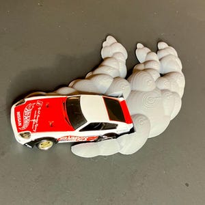 Puede incluir: Un coche de juguete blanco y rojo con las palabras "Hot Wheels" y "Nissan" en él, conduciendo a través de un objeto gris, texturizado, similar a una nube.