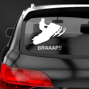 May include: White vinyl sticker of a snowmobile with a rider, with the text "BRAAAP!!" below. The sticker is on the back window of a black car.