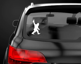 May include: White vinyl sticker of a Bigfoot silhouette giving the peace sign. The sticker is on the back window of a black car.